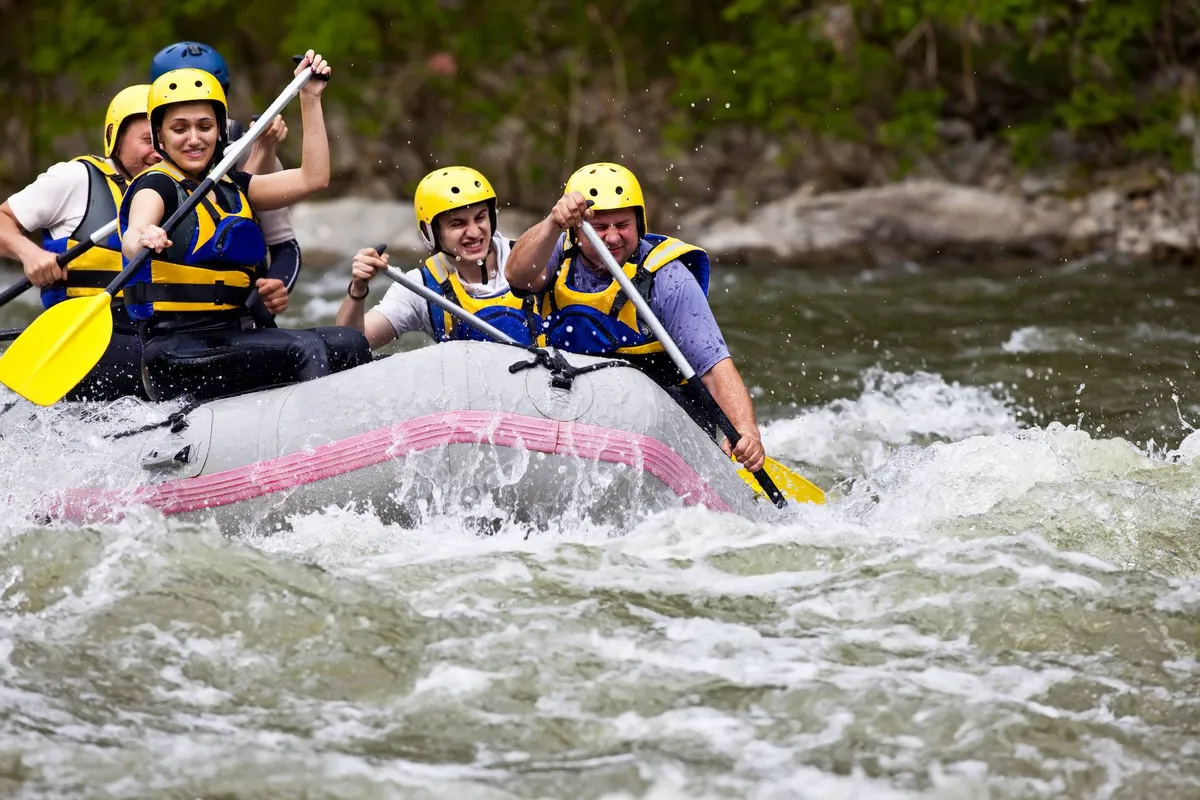 A group of people clad in life jackets maneuvers a raft through fast-moving river rapids, surrounded by lush, green, mountainous terrain. The outdoor setting is vibrant and energetic, reflecting both the excitement of adventure and the beauty of the region's natural landscape.