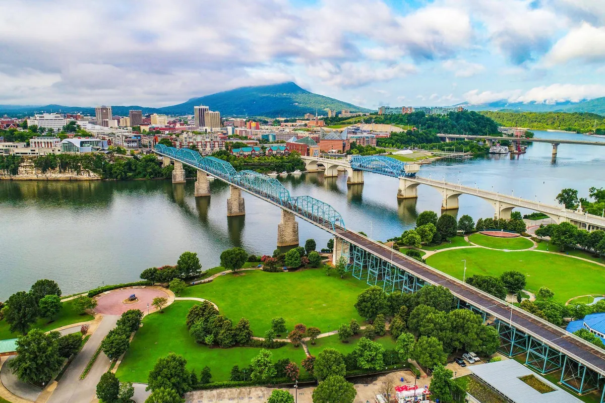 A broad view featuring the river, several city bridges, the Chattanooga skyline, and distant mountains. People are walking and biking on pathways along the water, suggesting easy access to both nature and urban amenities.