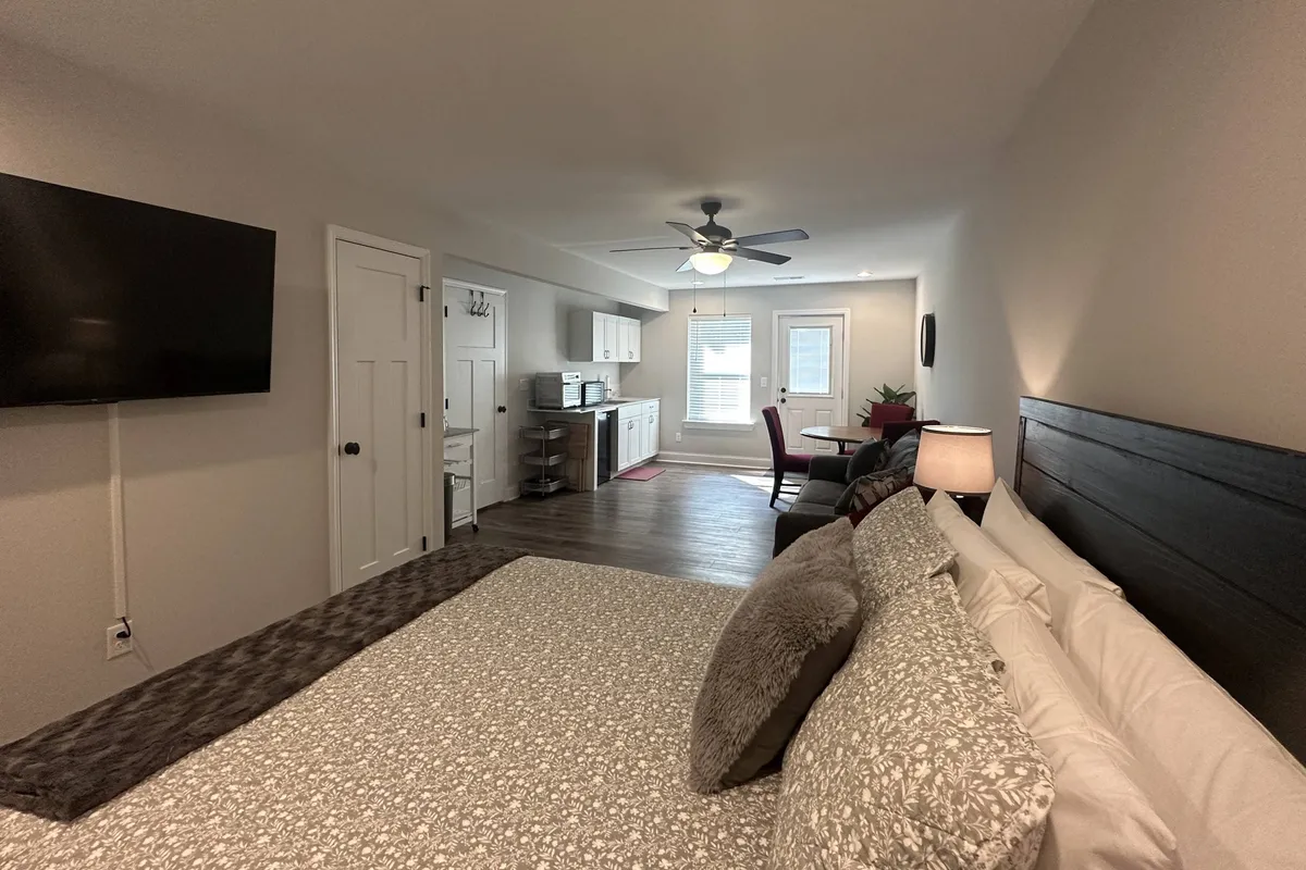 The hero image showcases a stylish, modern basement apartment with a plush king-sized bed dressed in inviting decorative pillows, a sleek flat-screen TV mounted on the wall, a compact yet fully equipped kitchen area with stainless appliances, and a cozy seating nook with a chair and table. The room is bathed in soft, natural light, creating a warm and peaceful ambiance ideal for unwinding after a day exploring Chattanooga.