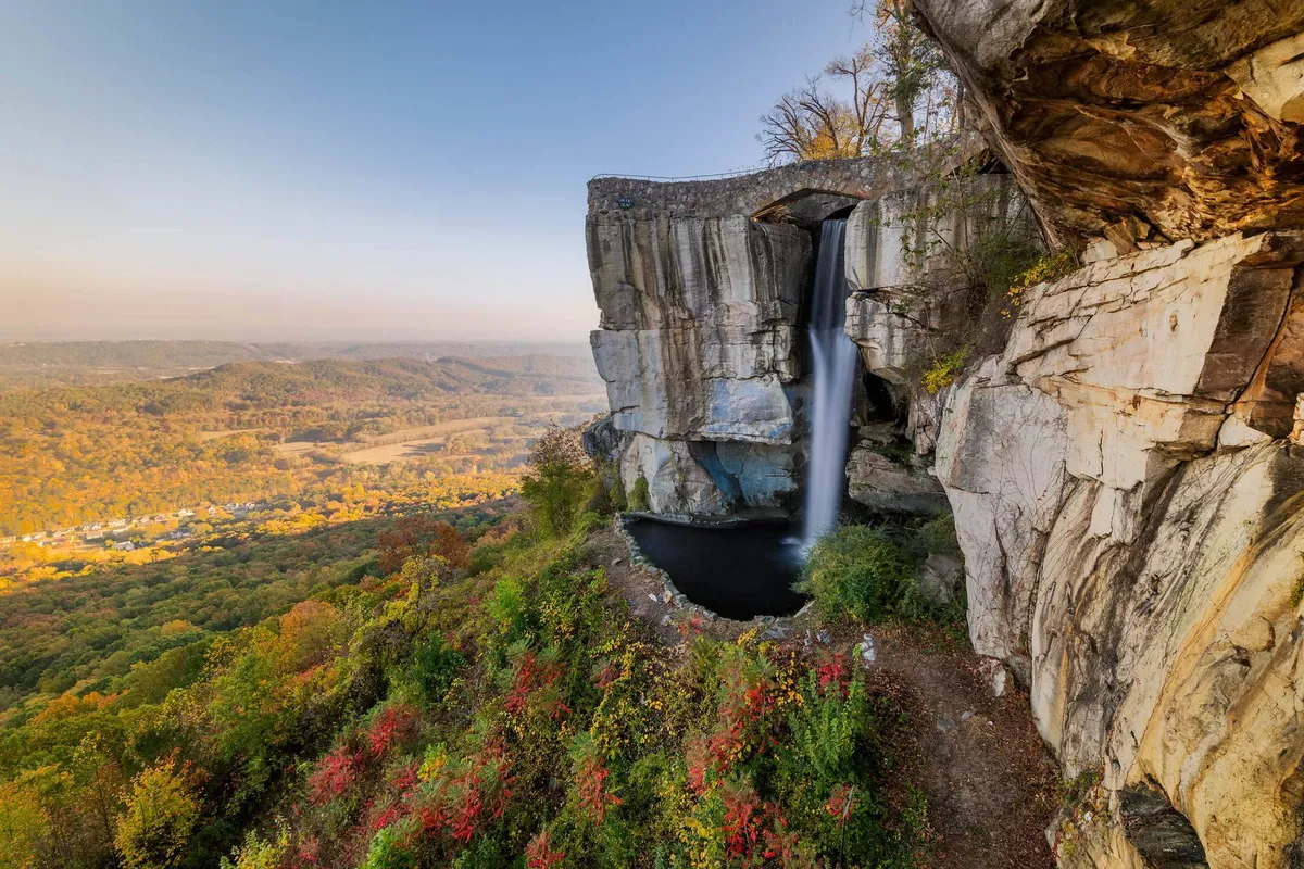 A scenic waterfall tumbles over rocks into a lush valley, framed by greenery and dramatic rock formations. The natural setting beckons for picnics, photography, and mindful relaxation.