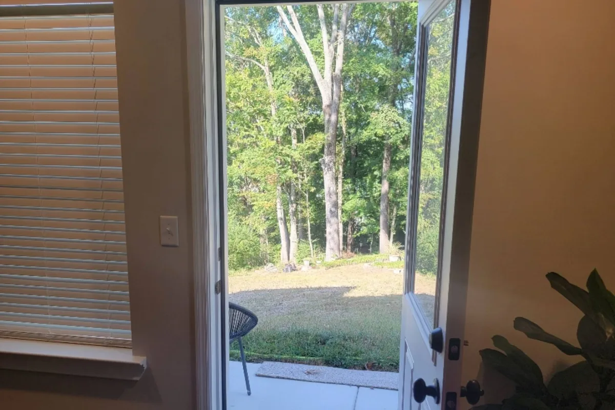 The image shows an open door leading from the apartment’s interior to a lush, green backyard. A comfortable chair sits on a patio, inviting residents to enjoy the fresh air. Morning light streams inside, emphasizing the seamless indoor-outdoor connection, while the landscape hints at the tranquility just steps away.