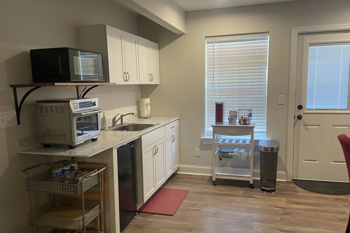 A cozy, functional kitchen scene featuring a modern countertop, sink, microwave, rolling cart, and a window with blinds. The space is well-lit and set up for both meal prep and enjoying coffee, highlighting the availability of quality appliances and thoughtful touches.