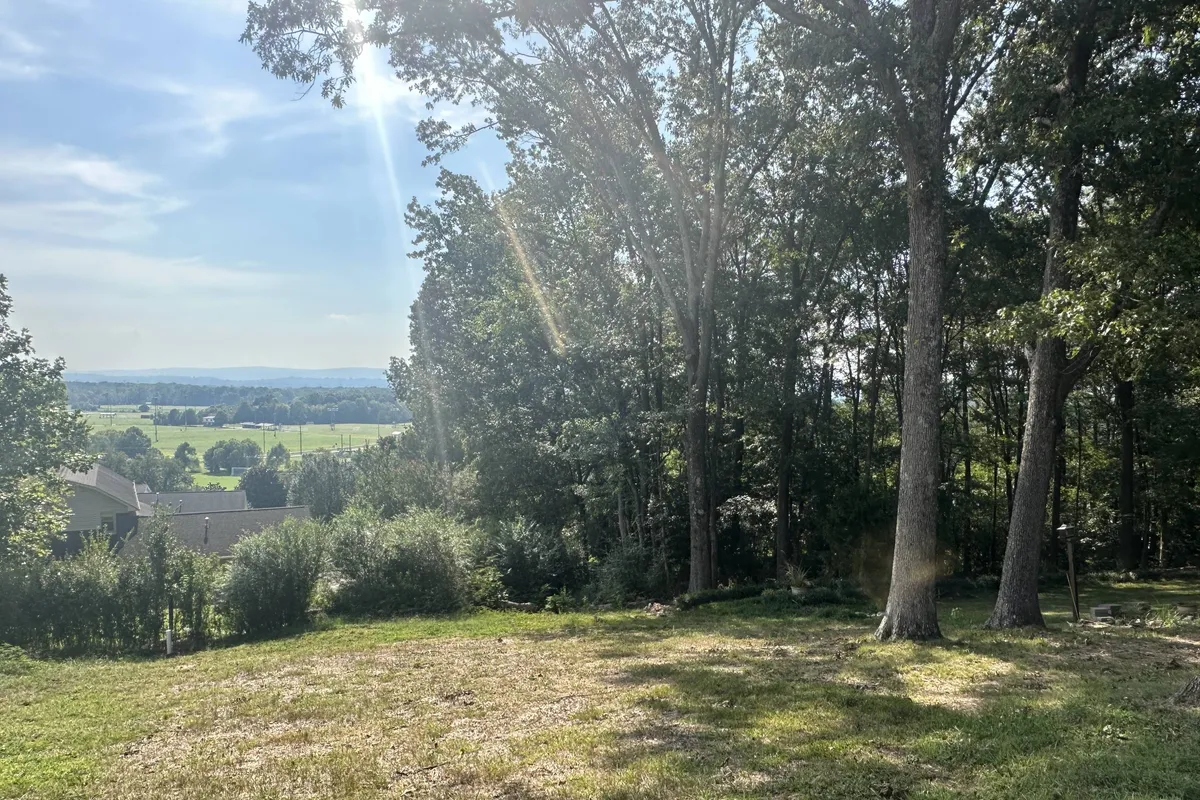 A sun-dappled landscape featuring lush greenery and distant hills, with sunlight streaming through the trees. The setting evokes tranquility and access to nature, showcasing the surrounding environment close to the property.