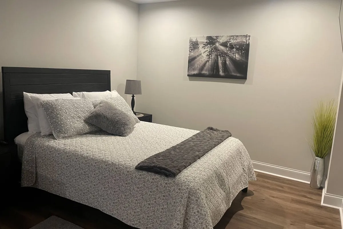 A bright, tranquil bedroom featuring a queen bed dressed in fresh linens, a decorative pillow, nightstand with lamp, wall art, and a leafy green plant. The styling is serene, cozy, and welcoming, with enough space to unpack and settle in comfortably.