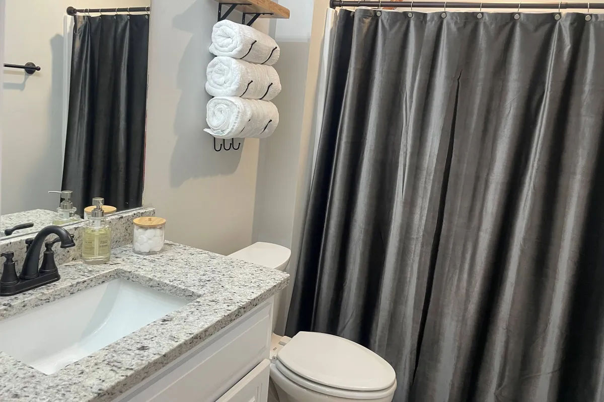 A stylish, modern bathroom with a granite countertop, black faucet, neatly stacked towels, and a sleek black shower curtain. The space is clean and functional, perfect for morning routines or unwinding after a long day.