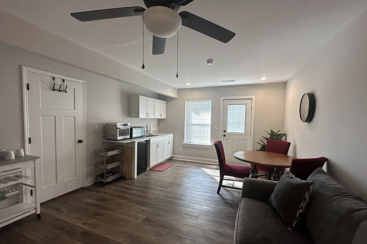 A bright, multifunctional living area featuring a sofa, a dining table, a kitchenette, and a doorway, all arranged in an inviting, homey layout. The scene conveys warmth, comfort, and the sense of a fully private living environment.