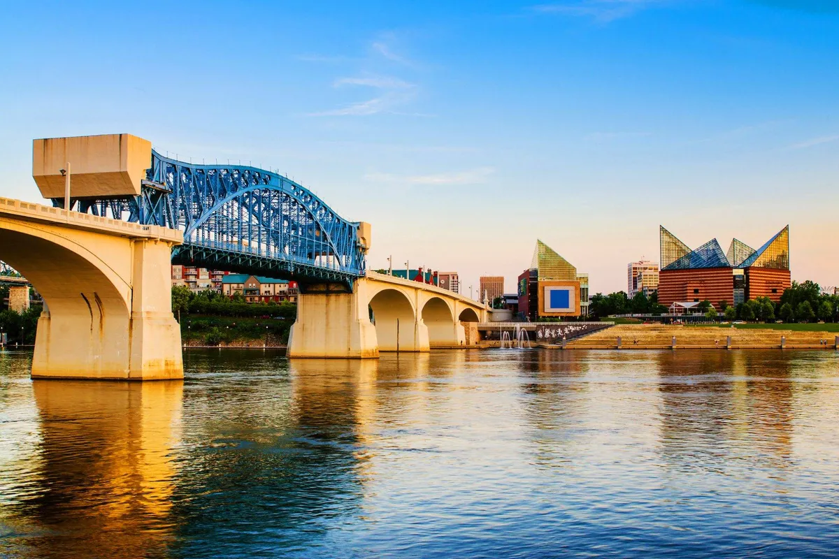 A scenic view of Chattanooga's iconic blue bridge stretching over the river, with modern downtown architecture in the background. The calm water captures reflections of the cityscape, and walkers and cyclists can be seen enjoying the sights along the waterfront.